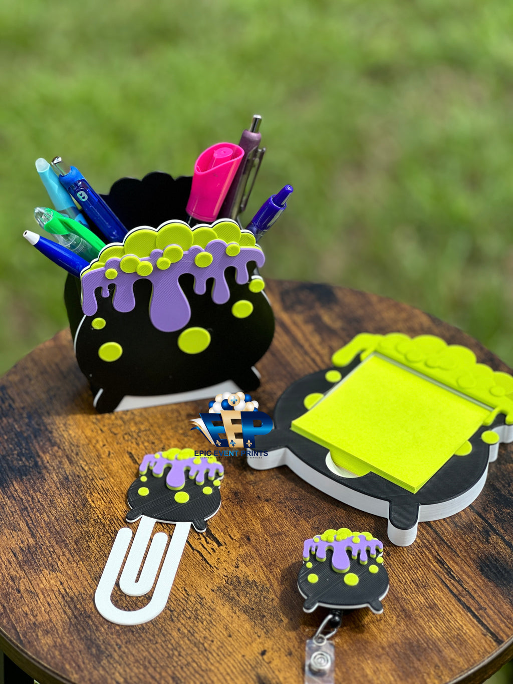 Four matching black cauldron desk accessories with purple drips and green bubbles: pen holder, sticky note holder, bookmark, and badge reel