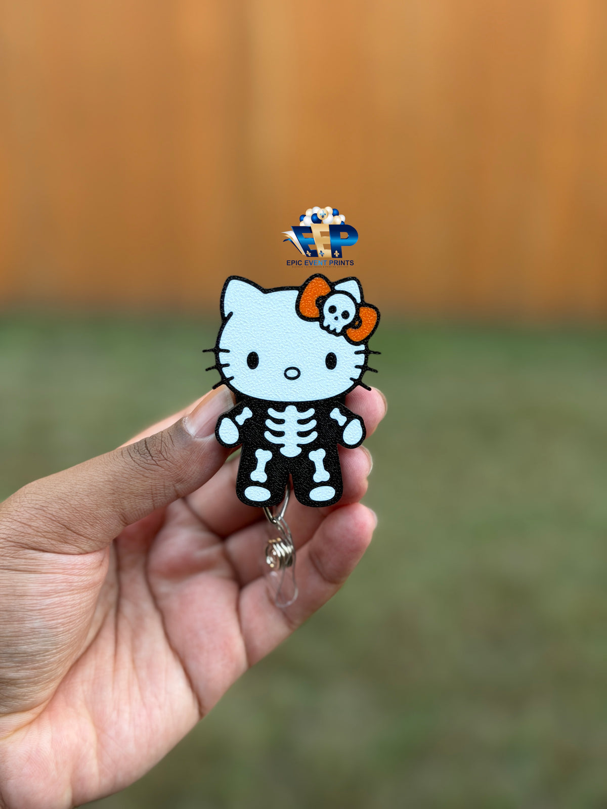 3D skeleton kitty badge reel with a white face, black-and-white skeleton outfit, and orange bow with skull detail, held in a hand.