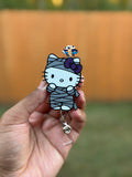 3D mummy kitty badge reel with a white face, gray mummy wraps, and a purple bow, held in a hand.
