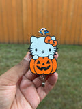 3D pumpkin kitty badge reel with a white kitty, orange bow, and orange jack-o’-lantern, held in a hand.