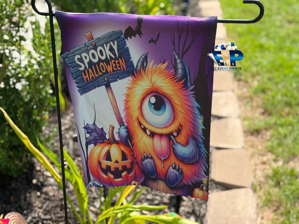 Purple one-eyed monster in witch hat under orange and purple spiderwebs on Halloween garden flag